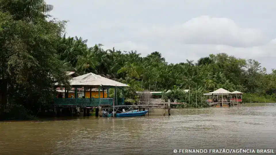 Comunidade ribeirinha às margens de um rio na Amazônia, com casas sobre palafitas e vegetação abundante, destacando a relação entre meio ambiente, clima e saúde.